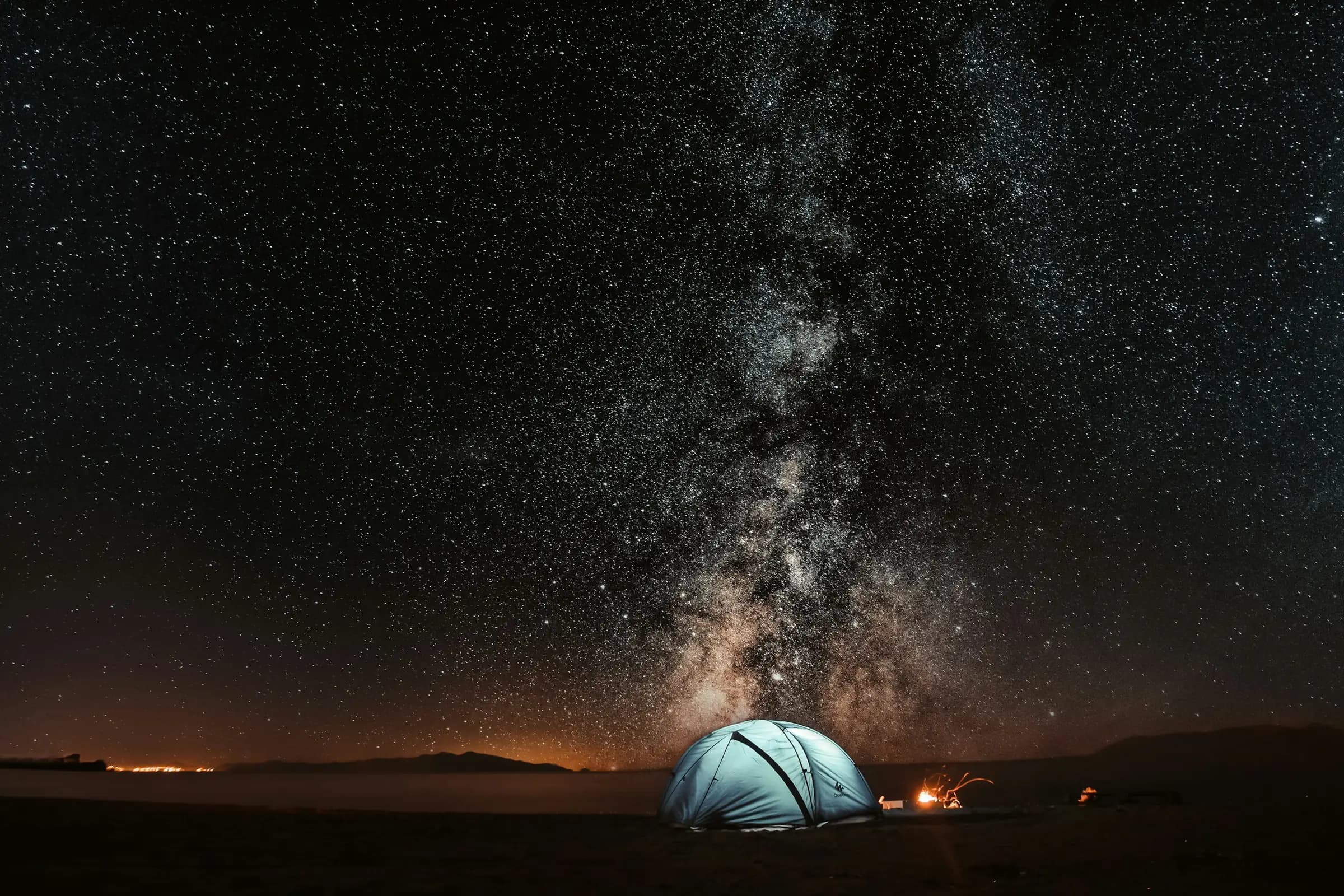 A lit tent glowing beside a campfire beneath the Milky Way at night.